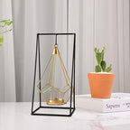 Geometric gold and black candle holder on a surface with books and a potted plant in the background.