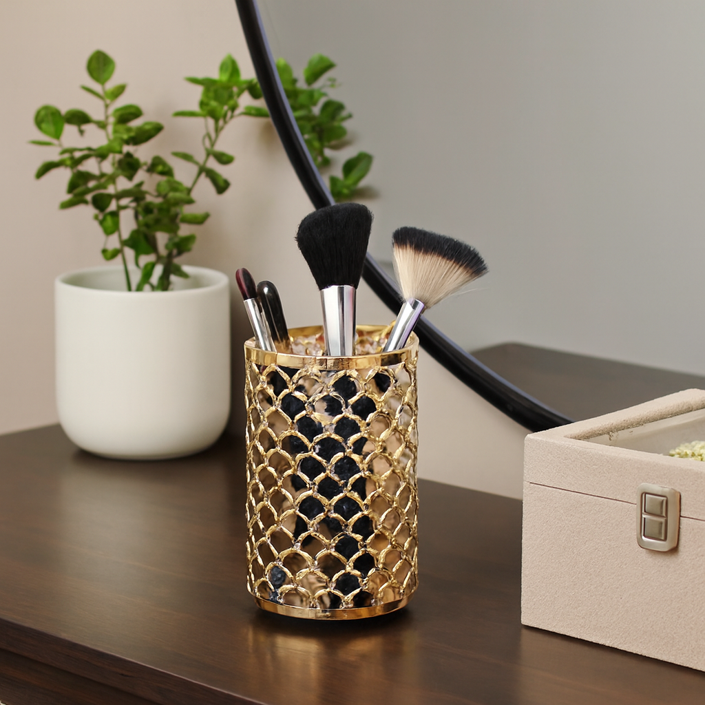 Gold decorative holder with makeup brushes on a vanity table with a plant and box in the background.