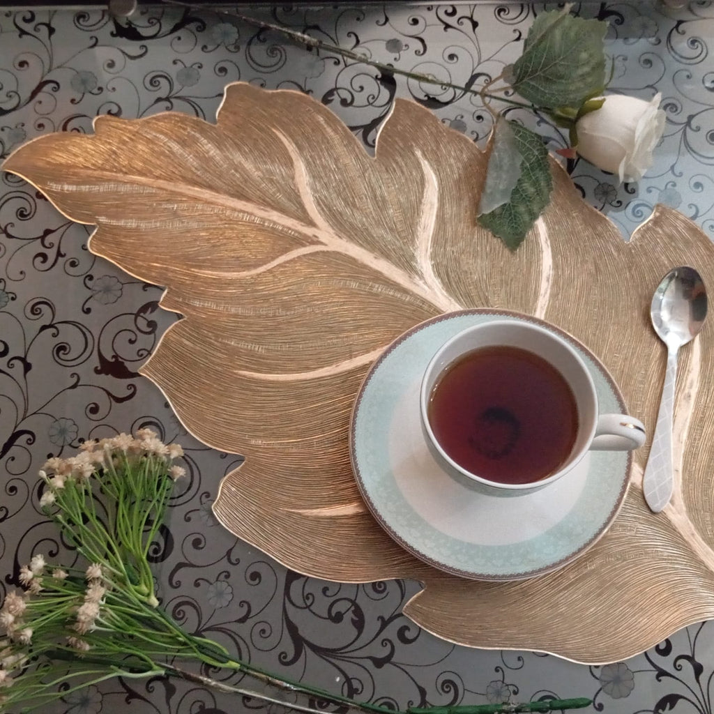 Tea cup on a decorative leaf-shaped platter with flowers and a spoon on a patterned tablecloth.