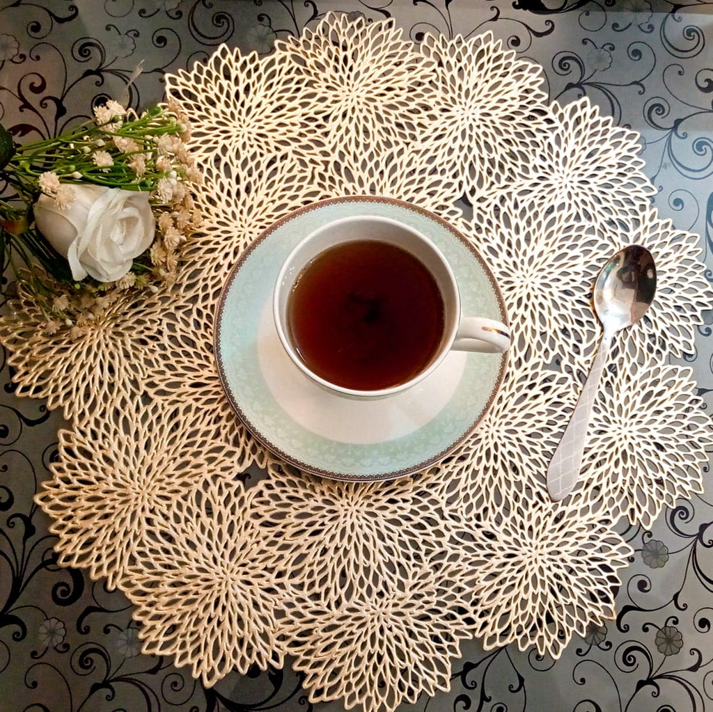 Tea cup and saucer on a decorative doily with a spoon and flowers on a patterned tablecloth.