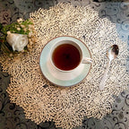 Tea cup and saucer on a decorative doily with a spoon and flowers on a patterned tablecloth.