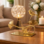 Decorative table lamp with crystal design and bird on a branch, placed on a wooden surface.