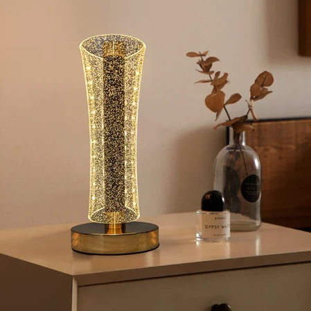 Decorative table lamp with a glass shade on a surface next to a bottle and a plant.
